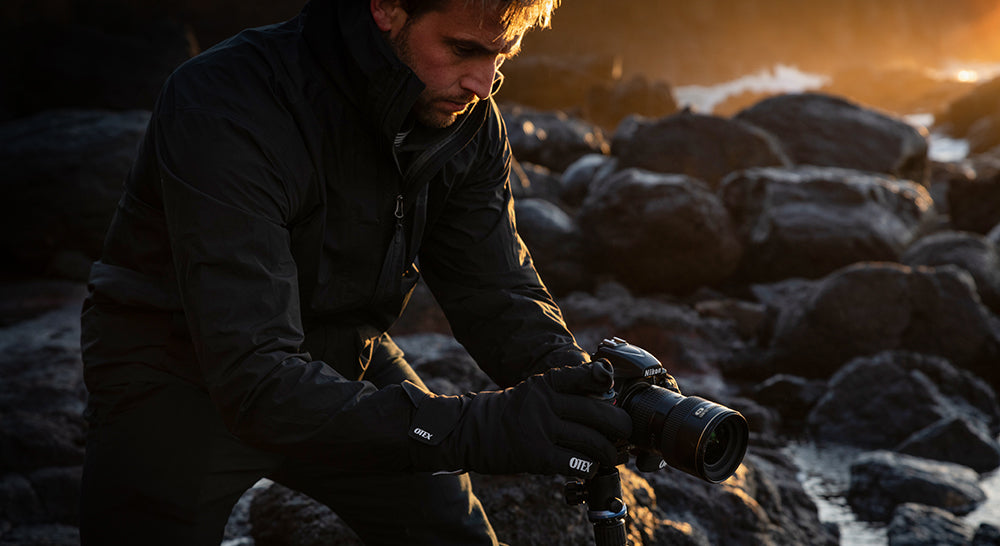 Person holding a camera wearing otex photography gloves on a rocky surface with a warm, golden light